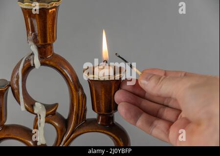 Die Hand eines Mannes hält ein erloschtes Streichholz gegen die brennenden Kerzen. Brauner Keramik-Kerzenständer im Hintergrund. Grauer Hintergrund. Nahaufnahme. Selektiv Stockfoto