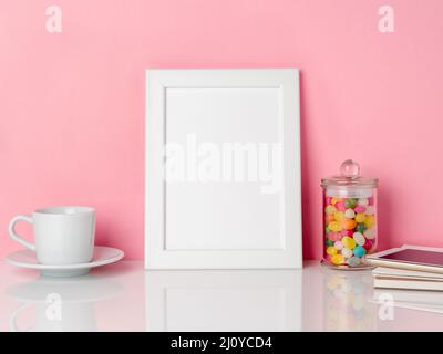 Leeren weißen Rahmen und Candys im Glas, Tasse Kaffee oder Tee auf einem weißen Tisch gegen die rosa Wand mit Kopie Raum Stockfoto