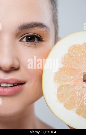Nahaufnahme der Grapefruit-Hälfte in der Nähe eines zugeschnittenen weiblichen Gesichts, isoliert auf Grau Stockfoto