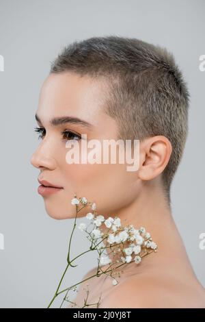 Porträt einer charmanten Frau mit kurzen Haaren in der Nähe von weißen winzigen Blumen isoliert auf grau Stockfoto