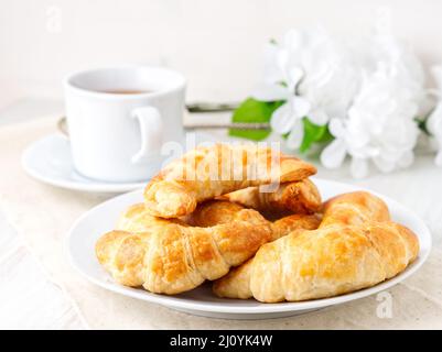 Tasse Tee, Blumen und frisch gebackenes Blätterteig auf einem weißen Tisch. Köstliche Croissants mit seitlichem Blick Stockfoto