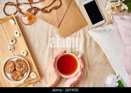 Frau, die einen Becher heißen Tees hält. Gemütlicher, gemütlicher Morgen im Bett. Flat Lay Woman Accessoires mit Brief, Umschlag, Smartphone, Parfüm. Stockfoto