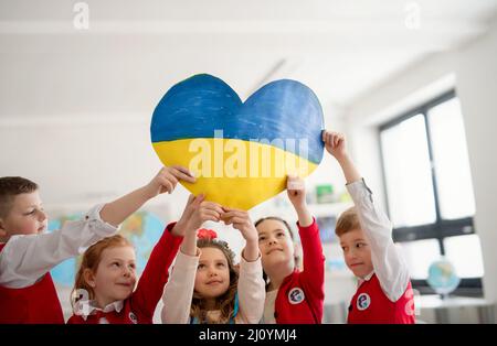 Kleine Schulkinder, die im Klassenzimmer ein Herz in ukrainischen Farben urlauben, kein Kriegskonzept. Stockfoto