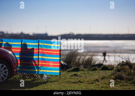 Eine Windpause am Barassie Beach in der Nähe von Troon , Ayrshire, Schottland, Großbritannien, zurück beleuchtet von Frühlingssonne, während ein entfernter, silhouettierter Mann auf dem Sand spaziert. Kredit: Windmill Images/Alamy Live Nachrichten Stockfoto