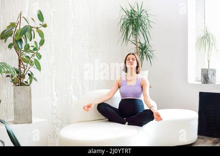 Fröhliche junge, schwanger Sportlerin, die in Lotuspose auf der Couch im hellen Raum sitzt und ihre Yoga-Session macht. Gesundheitswesen Stockfoto