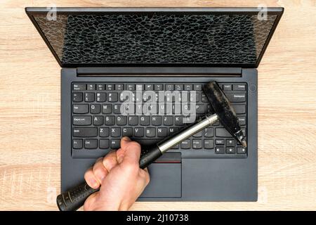 Hand mit einem Hammer und Laptop mit einem kaputten Bildschirm. Konzept der groben Reparatur der Computerausrüstung. Stockfoto