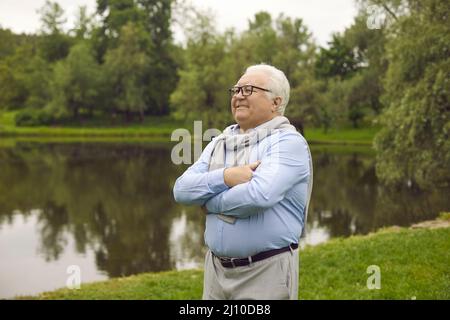 Glücklich lächelnd verträumter älterer Mann, der in der Nähe des Sees im Stadtpark nach vorne blickt Stockfoto
