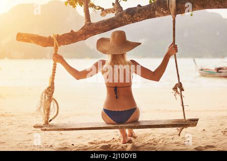 Sie schwingend sorgt sich weg. Rückansicht einer jungen Frau in einem Bikini, die auf einer Seilschaukel am Strand sitzt. Stockfoto