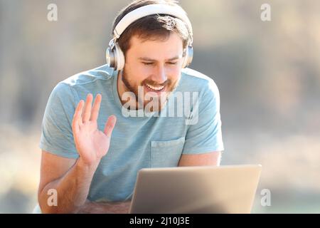 Fröhlicher Mann mit Kopfhörern, der während eines Videoanrufs auf einem Laptop im Freien mit der Hand begrüßt Stockfoto