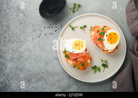 Sandwich mit leckeren Belägen geräucherter Lachs, Eier, Kräuter und Mikrogrüns Rettich, schwarze Sesam-Samen über weißem Teller auf grauem Beton Tisch Backgro Stockfoto