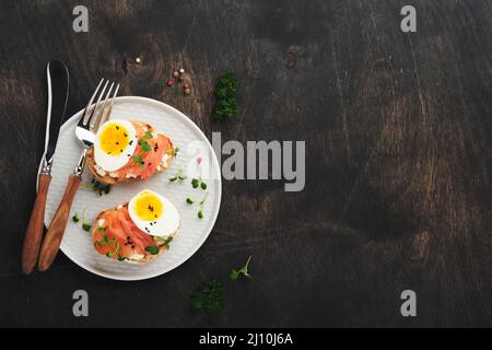 Sandwich mit leckeren Belägen geräucherter Lachs, Eier, Kräuter und Mikrogrüns Rettich, schwarze Sesam-Samen über weißem Teller auf altem Holztischhintergrund Stockfoto