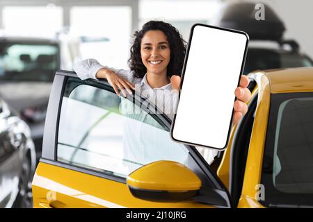 Positive Frau aus dem Nahen Osten, die mit dem Auto steht und das Smartphone zeigt Stockfoto