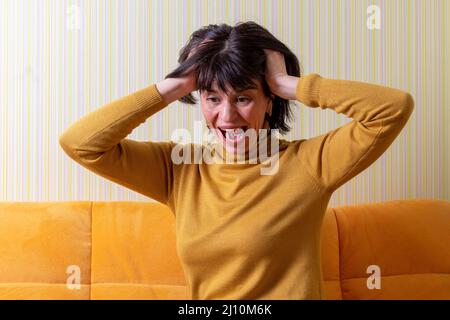 Verärgert schreiende Frau mittleren Alters, die unter Stress leidet und ihren Kopf mit den Händen auf einem orangefarbenen Sofa hält. Krise des mittleren Alters. Konzept der psychischen Gesundheit. Stockfoto