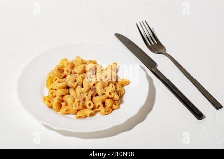 Lockige italienische Pasta mit Sauce auf einem weißen Teller. Minimalismus. Weißer Hintergrund. Pasta in Käsesauce. Messer und Gabel sind schwarz. Stockfoto