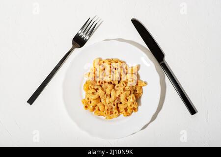 Lockige italienische Pasta mit Sauce auf einem weißen Teller. Minimalismus. Weißer Hintergrund. Pasta in Käsesauce. Messer und Gabel sind schwarz. Stockfoto