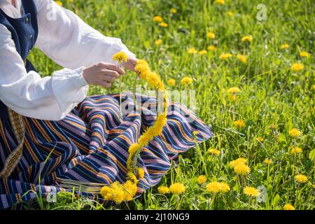 Junge Frau in Nationalkleidung mit gelbem Dandelion-Kranz im Frühlingsfeld. Frühling Stockfoto