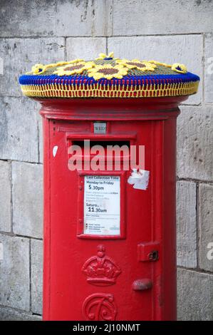 Ukrainische Farben blau und gelb: Gestrickter Wollhut ziert eine rote Kiste. In Solidarität mit dem Volk der Ukraine. Dorchester, Dorset, Großbritannien. Stockfoto