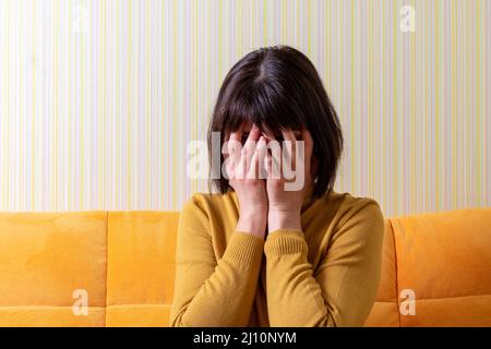 Aufgeregte weinende Frau mittleren Alters, die unter Stress leidet und ihr Gesicht mit den Händen auf einem orangefarbenen Sofa bedeckt. Krise des mittleren Alters. Konzept der psychischen Gesundheit. Stockfoto