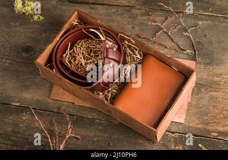 Handgefertigtes Geschenkset für einen Mann mit einem Ledergürtel und einem braunen Visitenkartenhalter in einer Schachtel auf einem alten Vintage-Holzhintergrund. Stockfoto