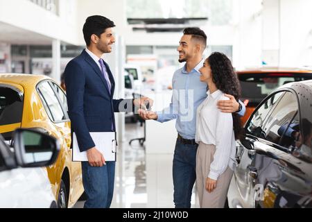 Fröhlicher Verkäufer, der dem arabischen Paar Schlüssel aus dem Auto gab Stockfoto