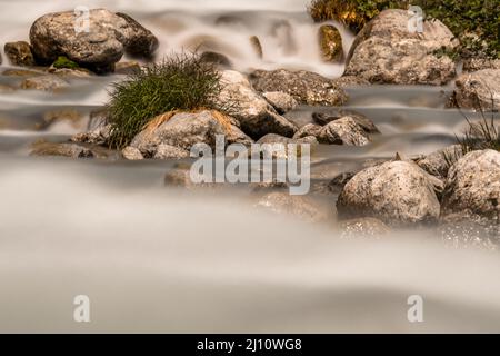 Lange Belichtung und Nahaufnahme eines Flusses, der zwischen Steinen und Grasklumpen fließt. . Hochwertige Fotos Stockfoto