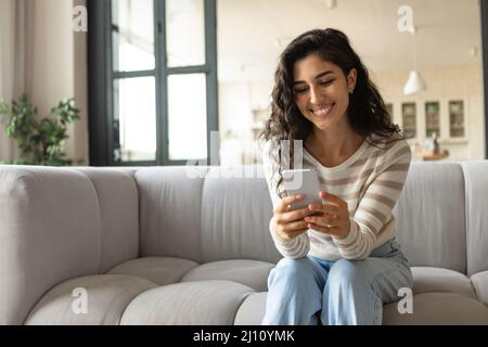 Tausendjährige kaukasische Frau mit Smartphone, während sie auf dem Sofa sitzt, Nachrichten überprüft, zu Hause Online-Videoanruf führt Stockfoto