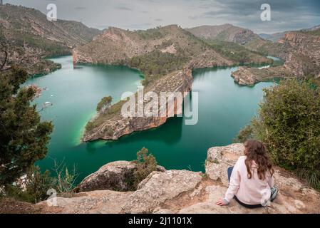 Junge Frau, die auf einem Felsen auf einem Berg sitzt, mit einer bergigen Landschaft und einem smaragdgrünen See im Hintergrund. Stockfoto