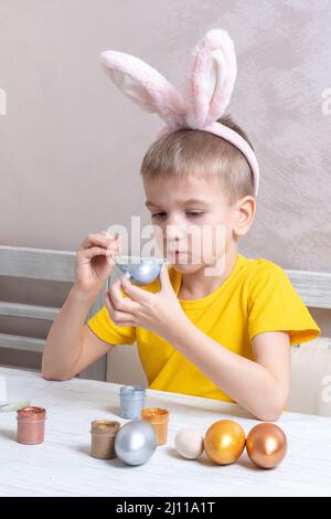 Ein kleiner blonder Junge in Hasenohren malt Eier für die Osterferien in der heimischen Küche, vertikaler Rahmen. Das Kind hat Spaß und feiert den Urlaub. Stockfoto