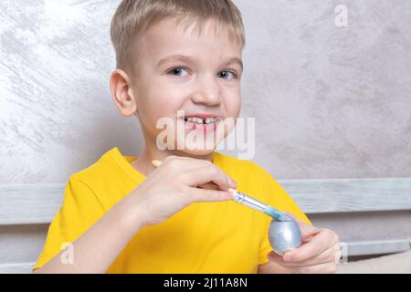 Ein kleiner blonder Junge malt Eier für die Osterferien zu Hause. Das Kind hat Spaß und feiert den Urlaub. DIY Ostereier Konzept. Stockfoto