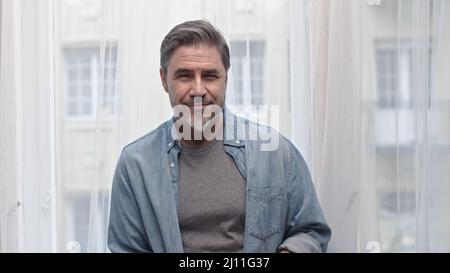 Porträt eines älteren grauhaarigen Mannes, der zu Hause im Fenster steht und lächelt. Stockfoto