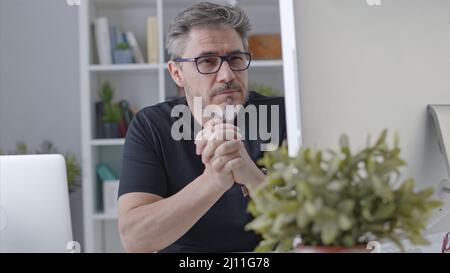 Ein älterer weißer Mann in einer Brille sitzt am Schreibtisch und benutzt im Heimbüro einen Desktop-Computer. 50s Geschäftsmann bleibt zu Hause in zwangloser Online-Arbeit Denken an Business-Lösung. Stockfoto