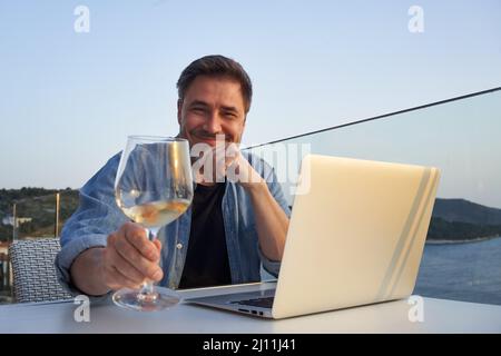 Geschäftsmann im Urlaub auf See, der mit einem Laptop arbeitet und Wein trinkt. Home Office, Online-Weinprobe, Telearbeit, Video-Chat. Flucht, Freiheit, Flucht . Stockfoto