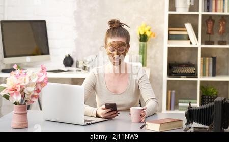 Frau kosmetische Gesichtshaut Pflege. Frau in der Haut Gummi Peeling Gesicht Schönheitsmaske sitzt am Schreibtisch mit Telefon und Laptop im Home Office. Stockfoto
