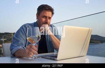 Geschäftsmann im Urlaub auf See, der mit einem Laptop arbeitet und Wein trinkt. Home Office, Online-Weinprobe, Telearbeit, Video-Chat. Flucht, Freiheit, Flucht . Stockfoto