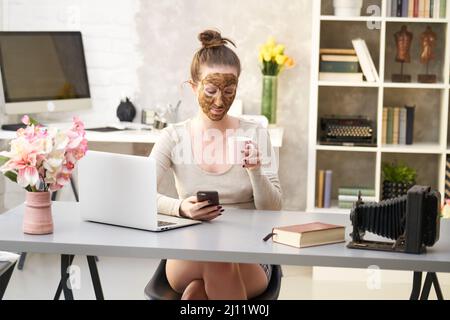 Frau kosmetische Gesichtshaut Pflege. Frau in der Haut Gummi Peeling Gesicht Schönheitsmaske sitzt am Schreibtisch mit Telefon und Laptop im Home Office. Stockfoto