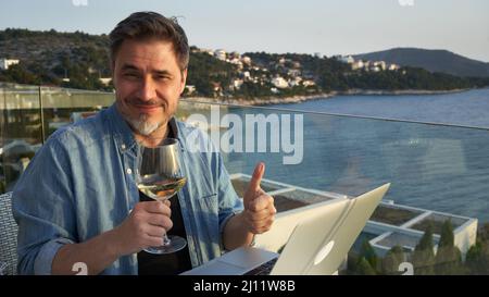 Geschäftsmann im Urlaub auf See, der mit einem Laptop arbeitet und Wein trinkt. Home Office, Online-Weinprobe, Telearbeit, Video-Chat. Flucht, Freiheit, Flucht . Stockfoto