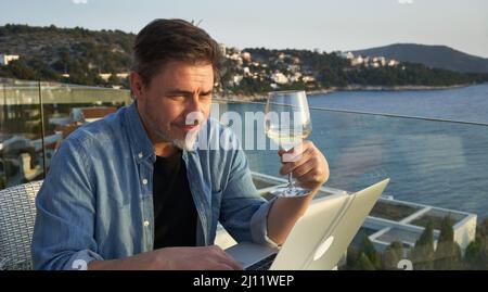 Geschäftsmann im Urlaub auf See, der mit einem Laptop arbeitet und Wein trinkt. Home Office, Online-Weinprobe, Telearbeit, Video-Chat. Flucht, Freiheit, Flucht . Stockfoto