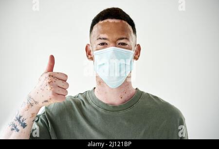 Seien Sie ein herausragender Bürger und üben Sie Sicherheit an erster Stelle. Studioporträt eines jungen Mannes mit Vitiligo, der eine Maske trägt und die Daumen gegen einen weißen zeigt Stockfoto