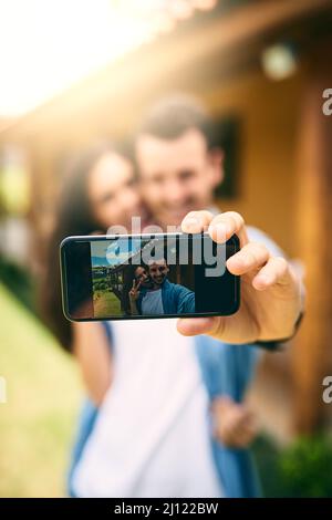 Ihre Liebe festhalten. Ausgeschnittene Aufnahme eines liebevollen jungen Paares, das draußen Selfies mit seinem Haus im Hintergrund macht. Stockfoto