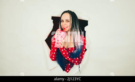 Barkeeperin mit Dreadlocks und einer Tasse süßem Getränk, die im Studio durch einen zerrissenen weißen Papierhintergrund guckt und auf die Kamera schaut. Stockfoto