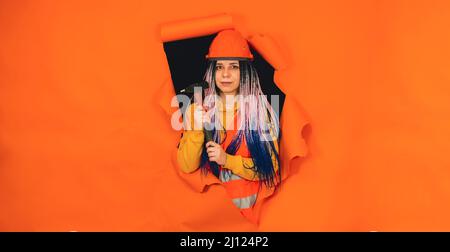 Bauarbeiterin mit Hammer, der aus dem Loch mit orangefarbenem Hintergrund herausragt. Junge Frau in Overalls mit Werkzeug und Blick auf die Kamera Stockfoto