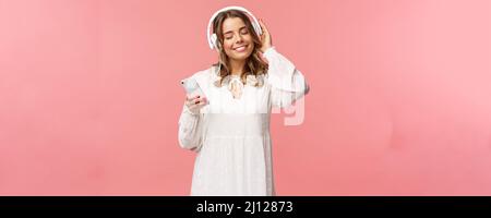 Porträt eines zärtlichen, fröhlichen und süßen blonden Mädchens in weißem Kleid, mit geschlossenen Augen und einem Lächeln, das sich wie Musik in Kopfhörern anhört, Wärme fühlt und Stockfoto