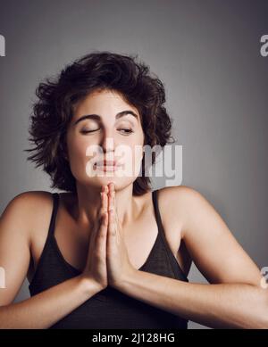 Immer noch ein bisschen Angst vor der Dunkelheit. Studioaufnahme einer attraktiven jungen Frau mit ihren Händen in Gebetshaltung vor grauem Hintergrund. Stockfoto