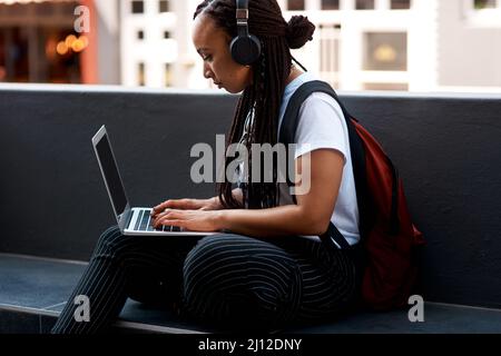 Verbinden Sie sich, machen Sie sich auf den Weg. Aufnahme einer attraktiven jungen Frau, die Musik hört und ihren Laptop benutzt, während sie sich im Freien entspannt. Stockfoto