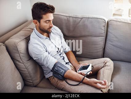 Ich mache meine regelmäßige Untersuchung. Aufnahme eines jungen Mannes, der zu Hause mit einem Blutdruckmessgerät seine eigenen Blutdruckwerte abnahm. Stockfoto