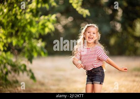 Glückliches kleines blondes Mädchen, das mit Kopfhörern Musik hört, in der Natur im Park tanzt und singt. Stockfoto