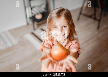 Ein kleines süßes blondes Mädchen mit langen Haaren lacht fröhlich über die Kamera und hält einen Apfel in den Händen Stockfoto