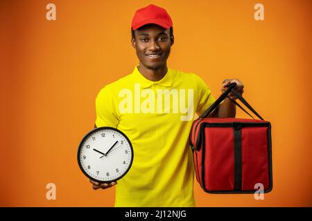 Lächelnder afroamerikanischer Kurier mit Uhr und Box auf orangefarbenem Hintergrund Stockfoto
