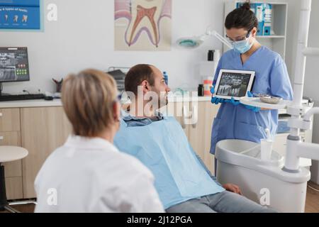 Stomatologe Krankenschwester hält Tablet-Computer erklären Radiographie Expertise Diskussion Zahnbehandlung mit dem Patienten während der Stomatologie Beratung in der Zahnarztpraxis. Ärzteteam trägt Gesichtsmaske Stockfoto