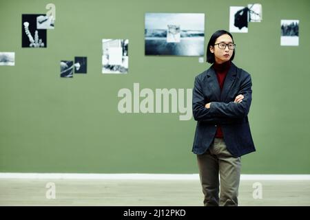 Horizontales mittellange Porträt eines jungen asiatischen Mannes mit Brillen, die mit gekreuzten Armen in der Galerie für moderne Kunst stehen und Fotos in der Ausstellung betrachten Stockfoto